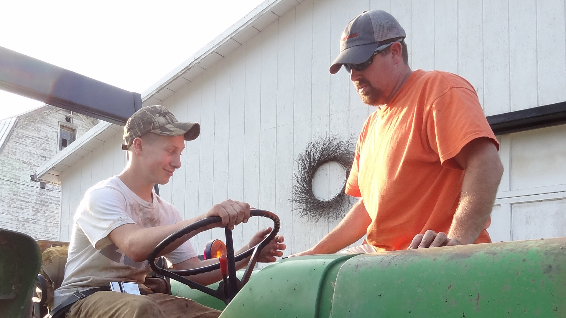 Two individuals talking while seated and standing on a green tractor near a farm building.